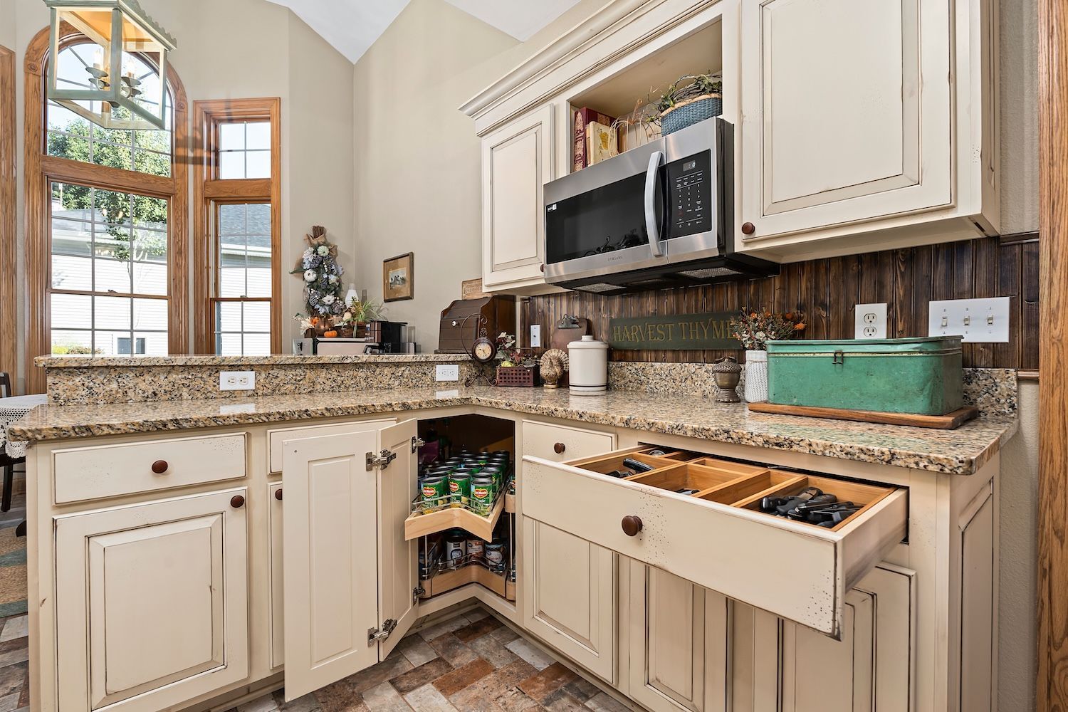 Cream-colored kitchen with granite countertops, open cabinet revealing soda, and silverware drawer, near a large arched window.