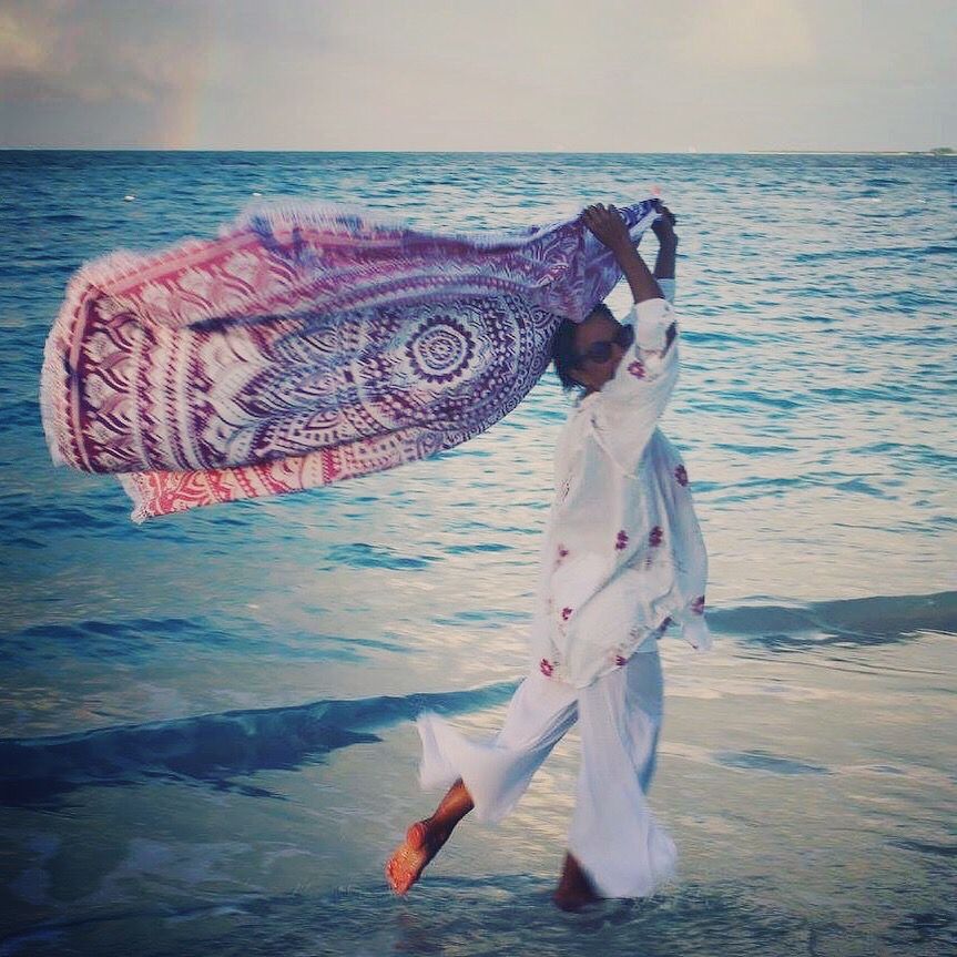A woman in a white dress is standing on the beach holding a scarf