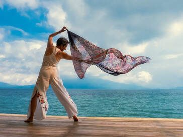 A woman is standing on a pier holding a scarf in front of the ocean.