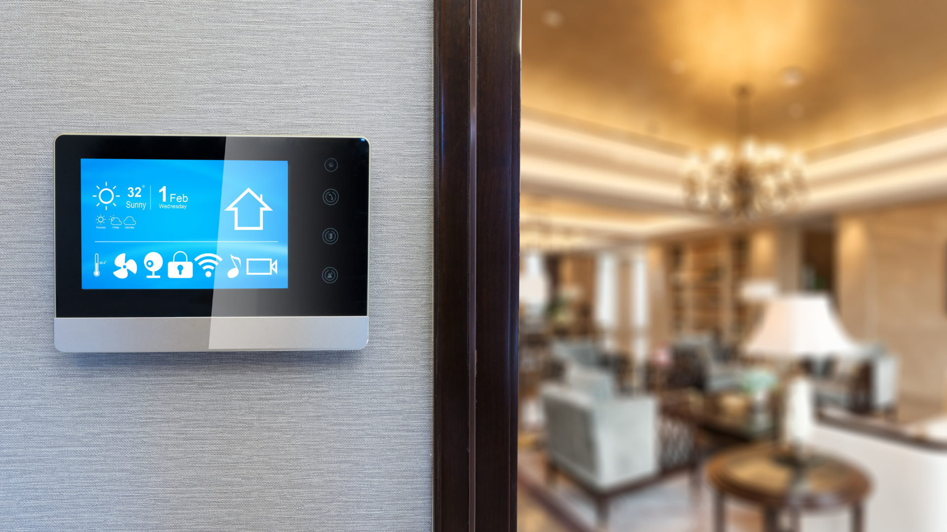 Smart home control panel on a wall, displaying icons and temperature. Interior with blurred living room view.