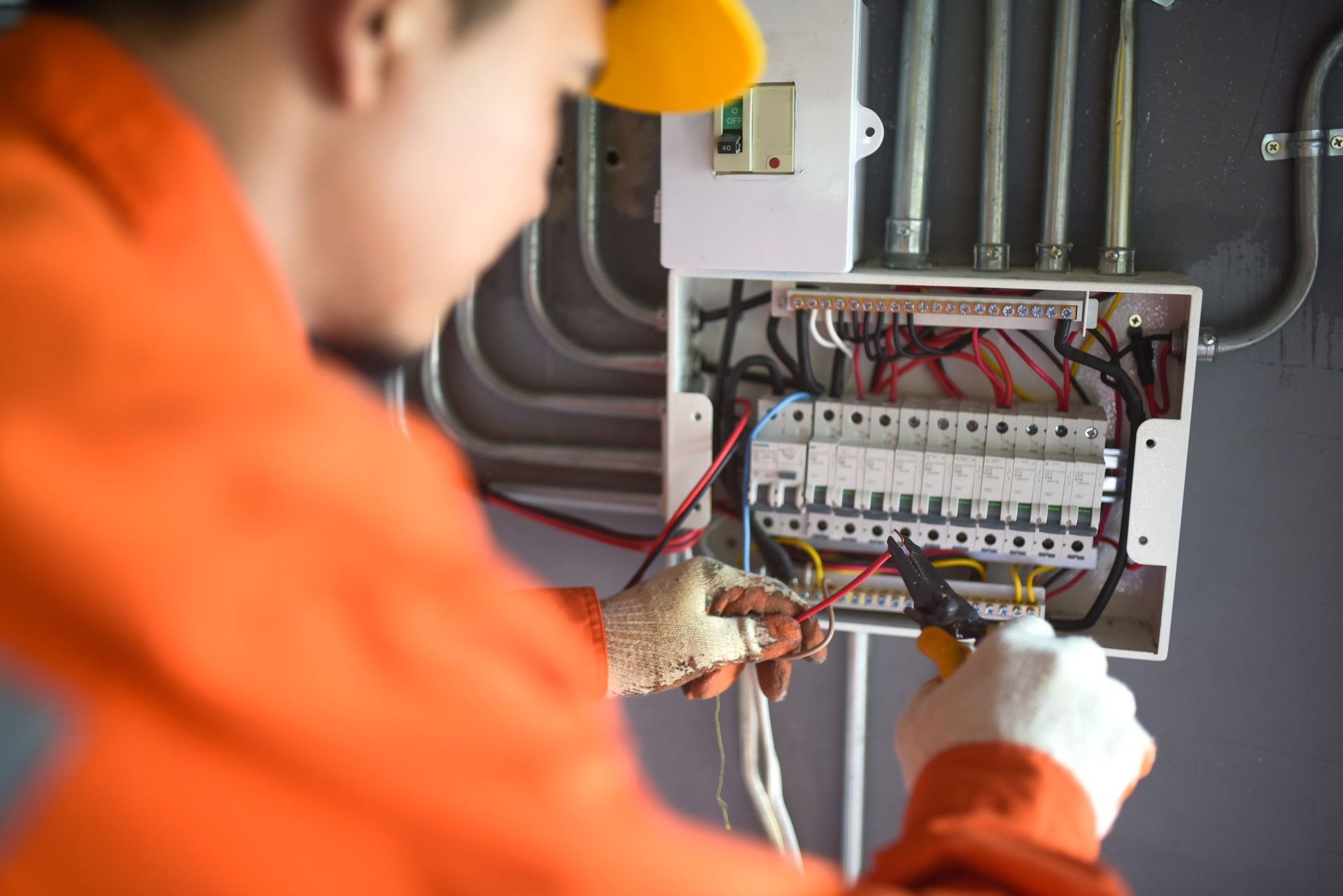 An electrician in an orange uniform is repairing a circuit breaker panel, holding wires with pliers.
