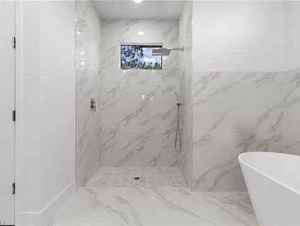 Amplio baño con ducha y bañera con azulejos de mármol, paredes blancas y una ventana.