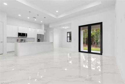 Cocina y sala de estar modernas y luminosas con gabinetes blancos, pisos con apariencia de mármol y grandes puertas con marco negro.