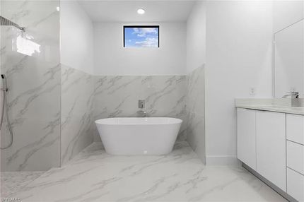 Baño blanco moderno con bañera independiente, azulejos de mármol y una pequeña ventana con vista al cielo.