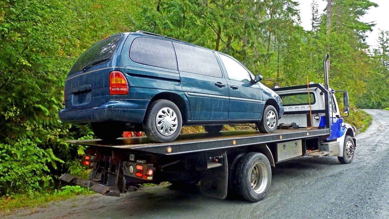 Blue minivan on a tow truck, on a gravel road next to trees.