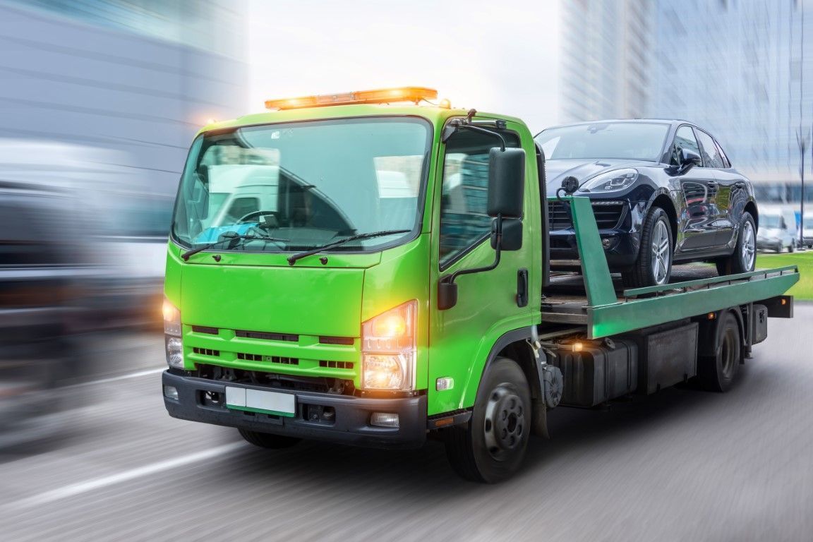Green tow truck carrying a black car on a city street with blurred background.