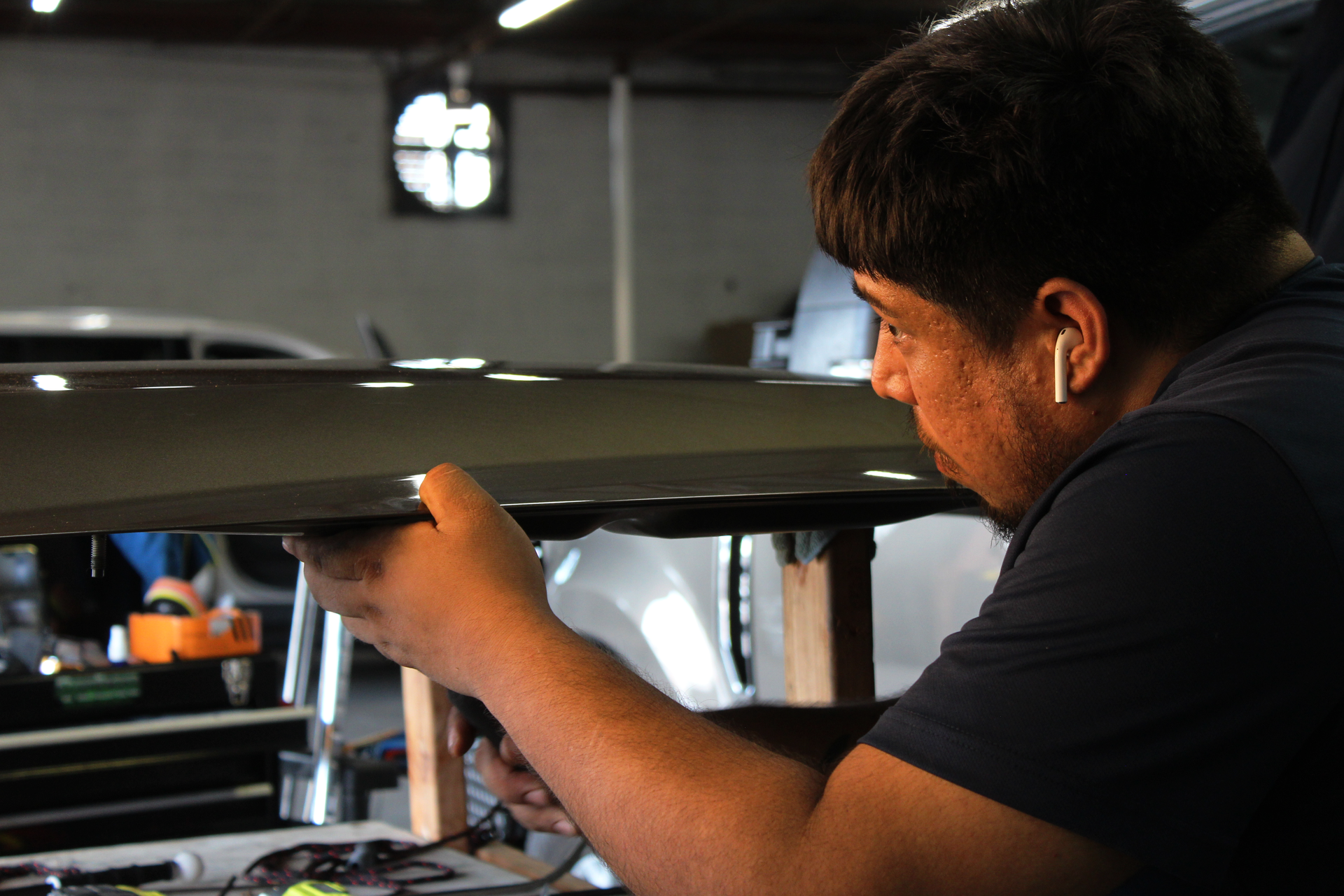 Man working on a car panel inside a garage, holding it and focused. Wearing earbuds.