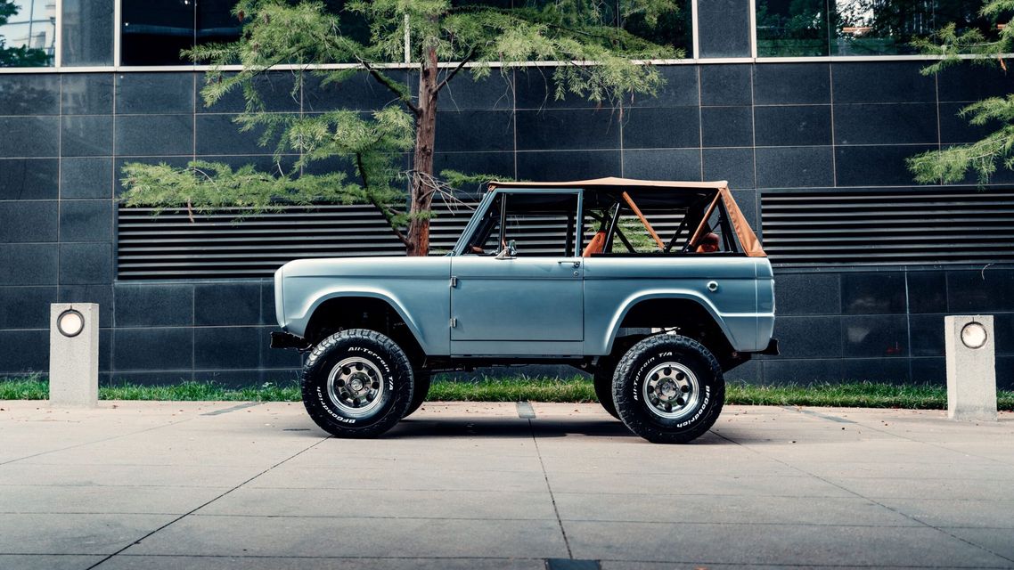 Light blue vintage Ford Bronco with a tan roll cage parked on a paved area next to a building.