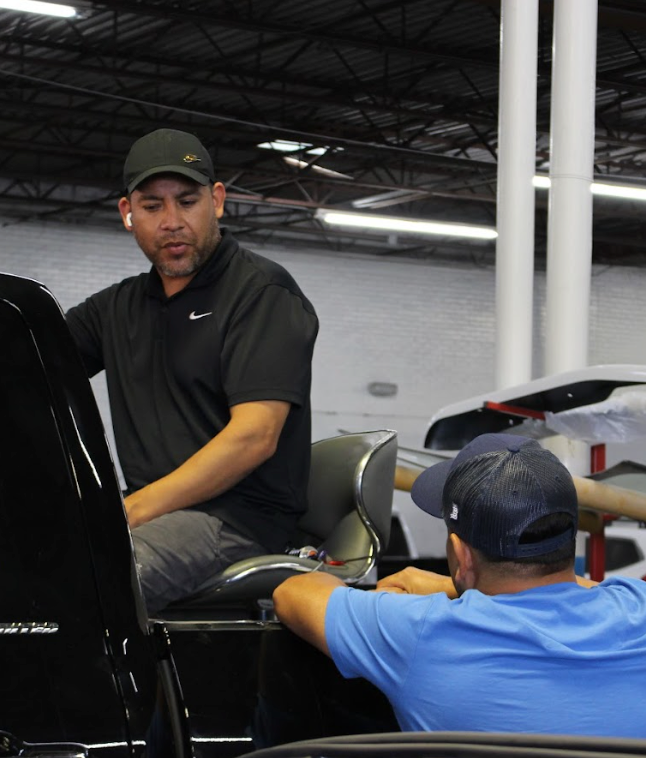 Two men working on a car in a garage. One wears a black shirt, the other a blue shirt.