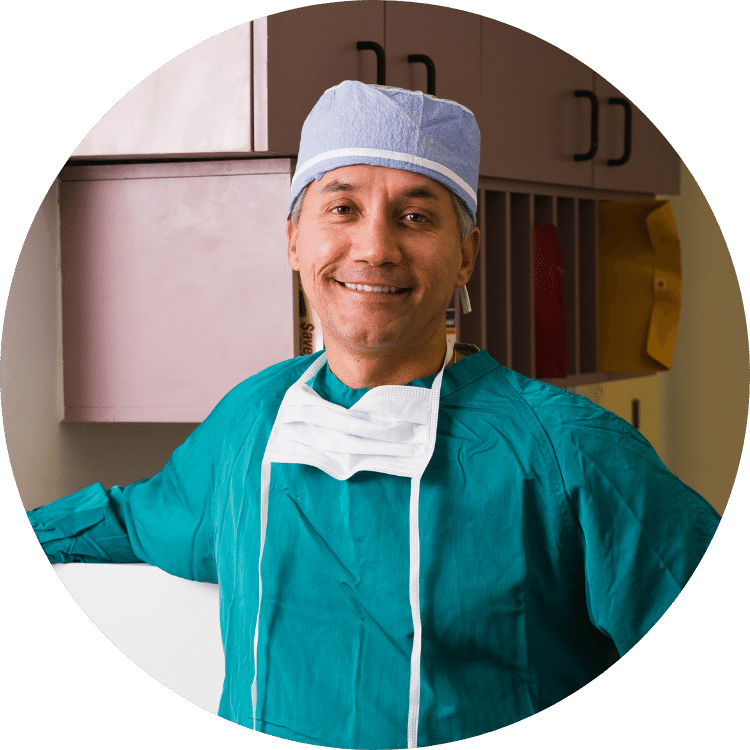 A surgeon wearing a surgical gown and mask smiles for the camera