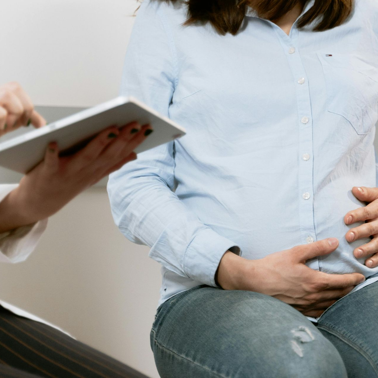 Pregnant woman seated, touching her abdomen. Another person holds a tablet nearby.