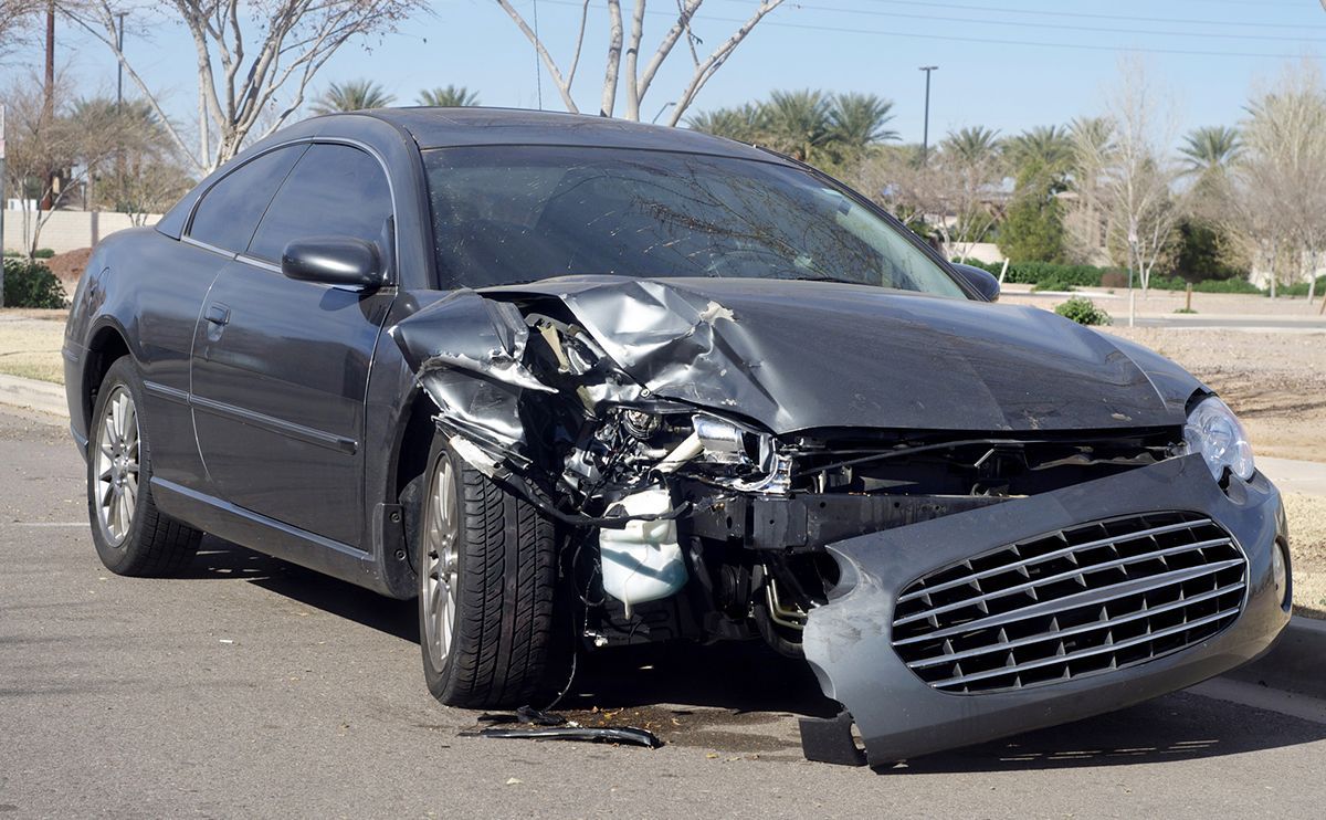 Damaged gray car with front-end collision on a paved road; surrounding trees and buildings.
