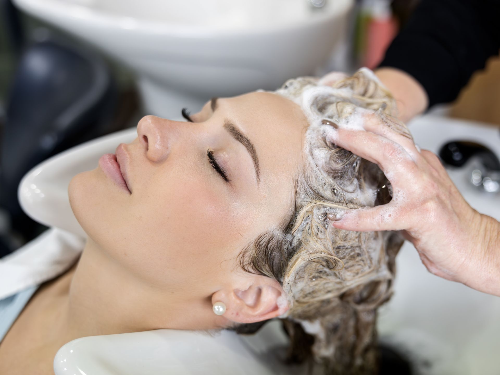 A person with eyes closed leans back in a salon sink as hands massage soapy lather into their hair.