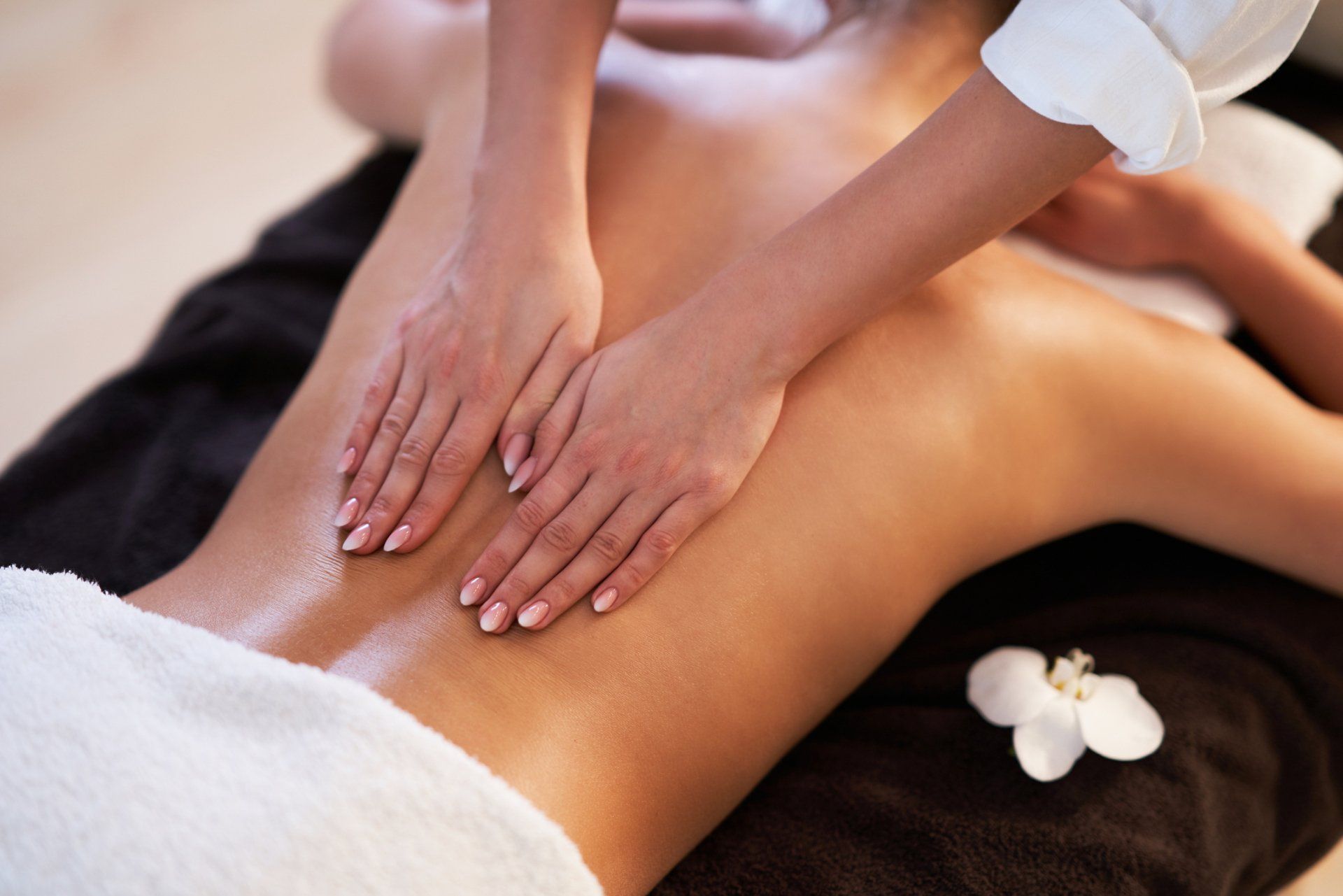 Hands performing a massage on a person's oiled, bare back with a white flower on a black towel nearby.