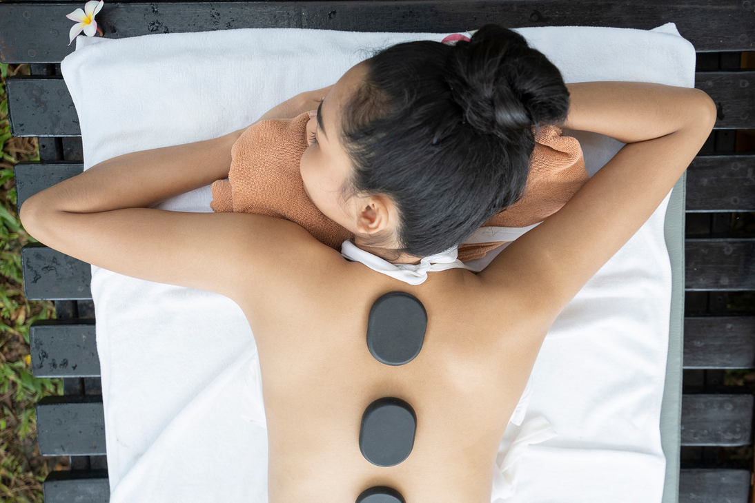 A person lying face down on a massage table receiving a hot stone treatment on their back.