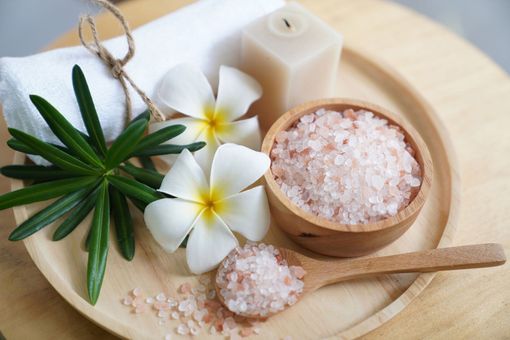 A wooden tray holding pink Himalayan salt in a bowl and spoon, white plumeria flowers, a towel, and a candle.