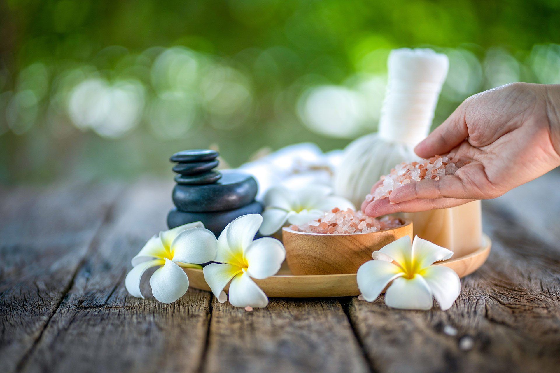 A hand holds pink Himalayan salt near spa elements: a stack of stones, plumeria flowers, and a herbal compress on wood.