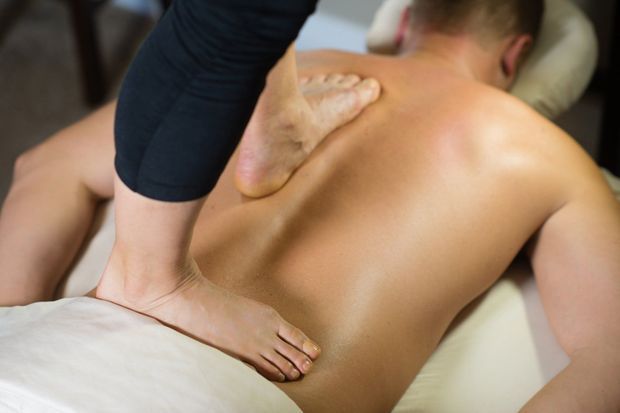 A massage therapist uses their feet to apply pressure to a person's back while they lie on a massage table.