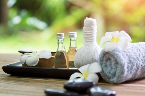 Spa items on a wooden tray: two glass bottles of oil, a white massage compress, a rolled towel, and frangipani flowers.