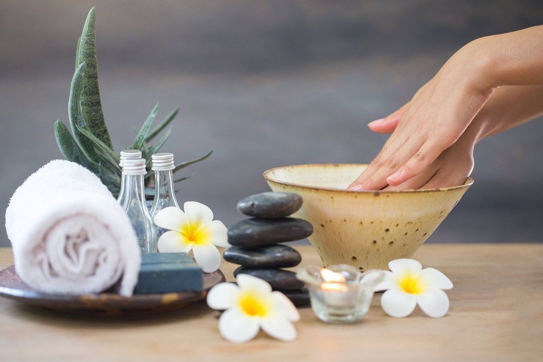 Hands soak in a spa bowl next to aloe, rolled towel, stones, and plumeria flowers on a wooden table.