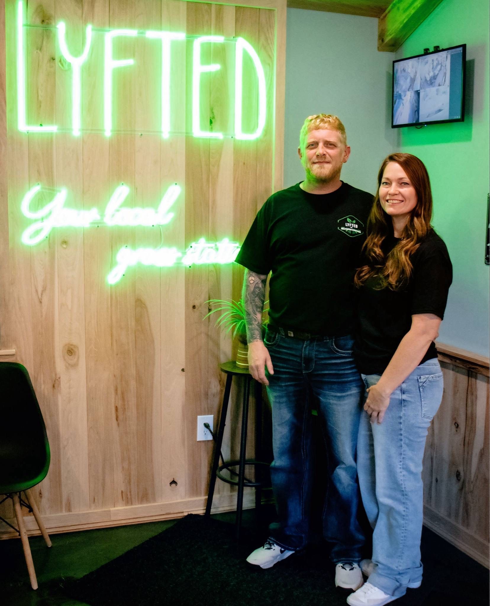 A man and woman stand together in front of a wood-paneled wall with a glowing green neon sign reading 