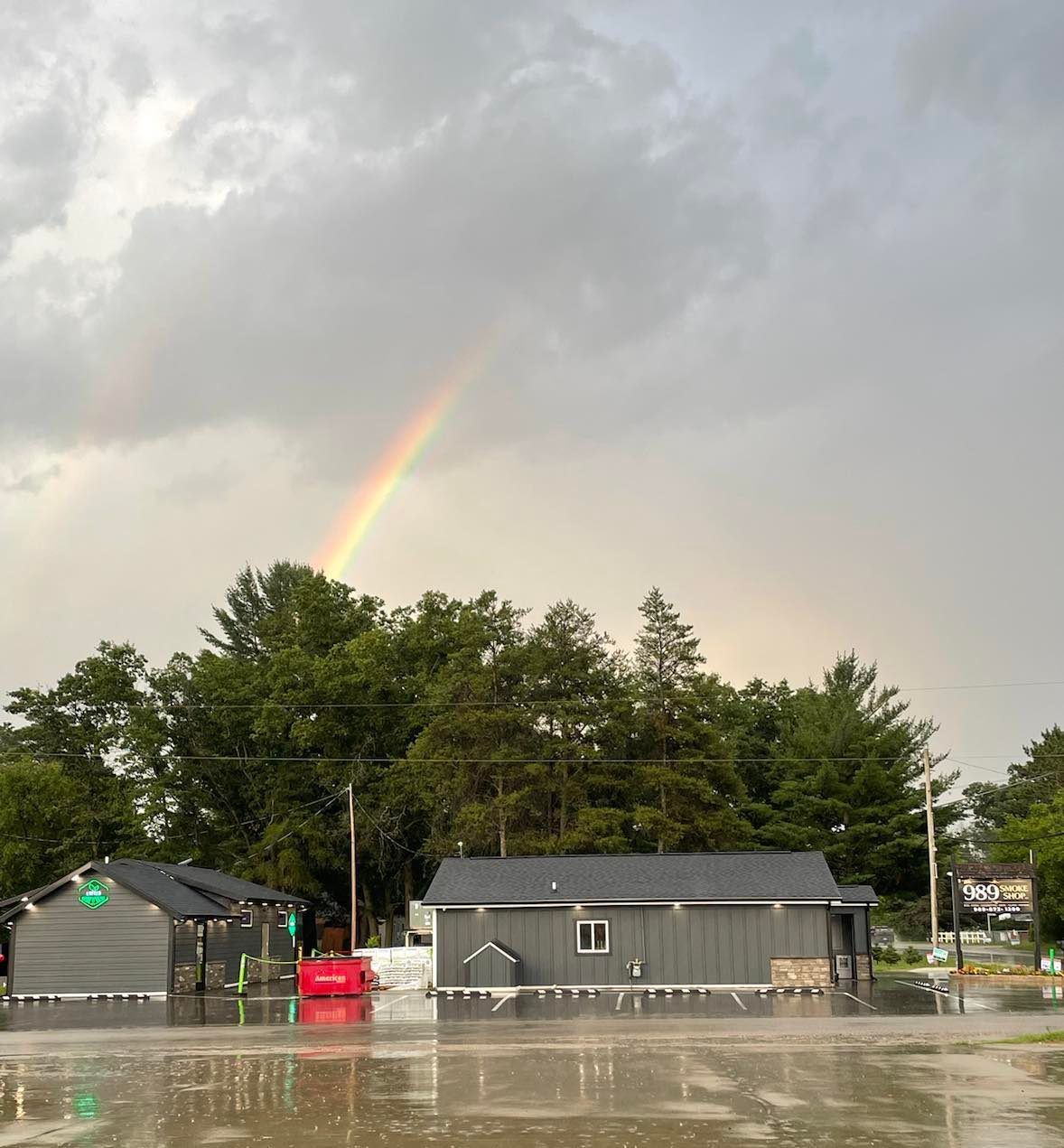 A faint rainbow arching over dark grey buildings and lush green trees in a rain-slicked parking lot.