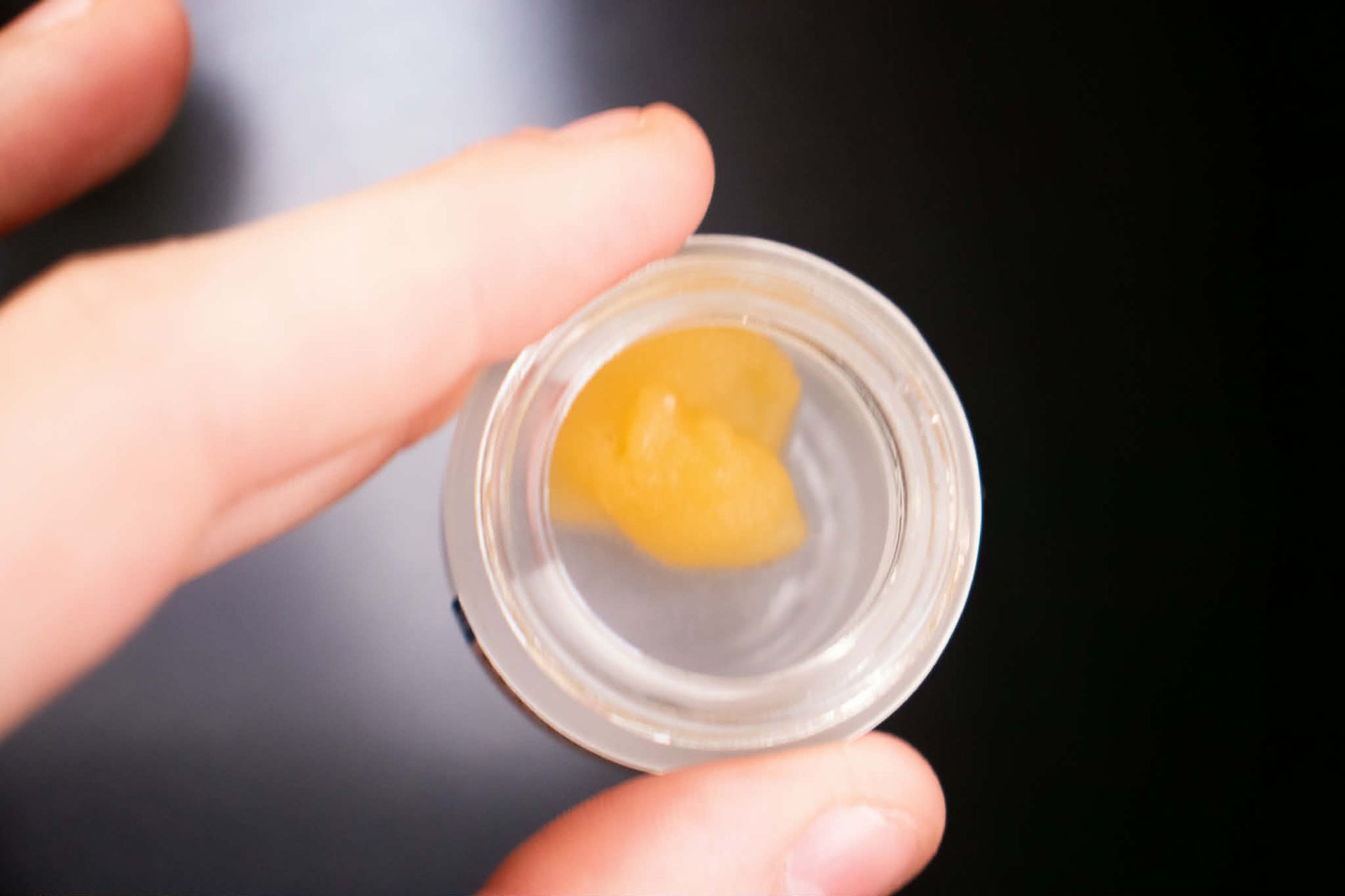 A hand holds a small glass jar containing a golden, waxy cannabis concentrate against a plain black background.