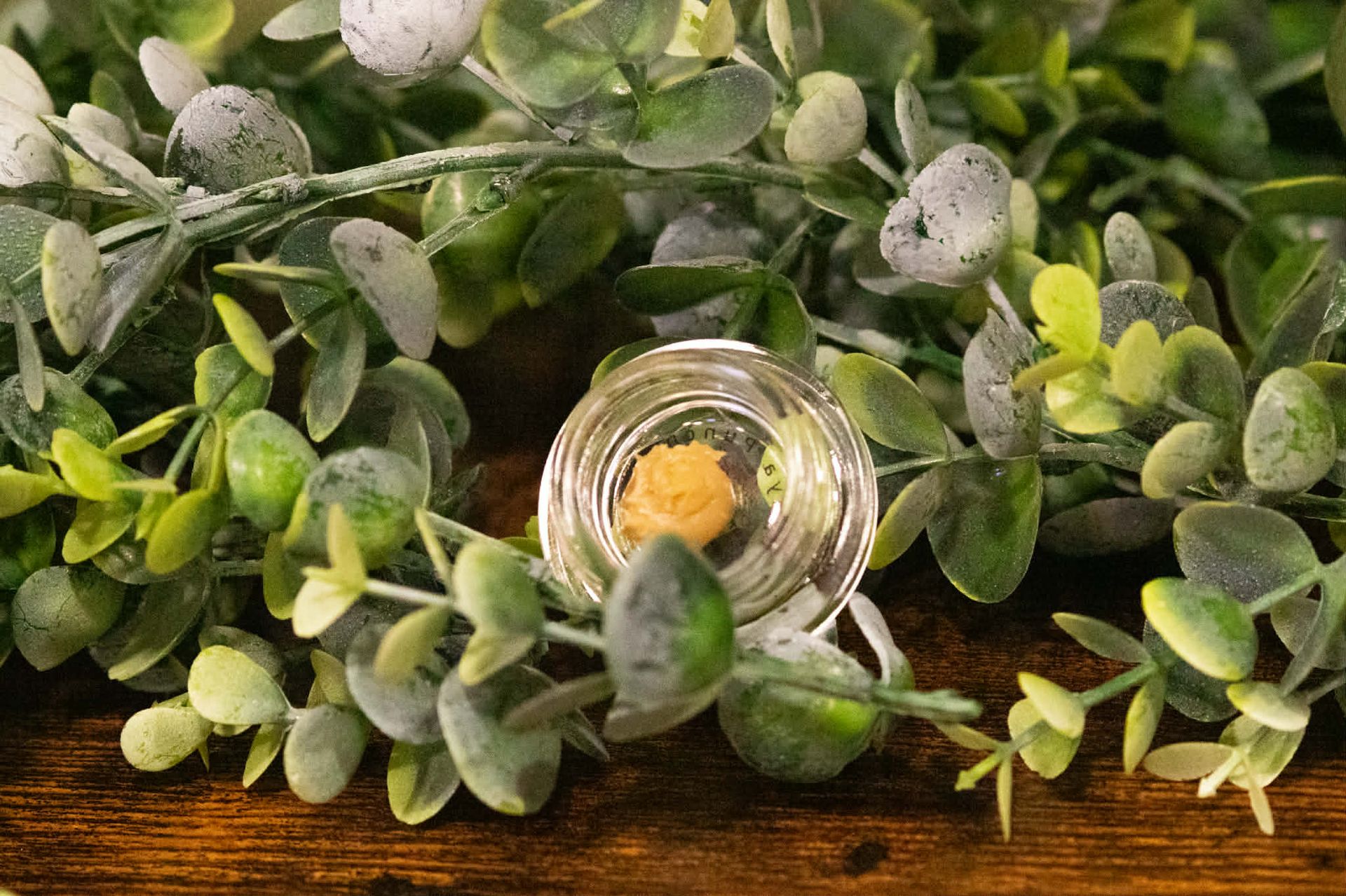 A small, clear glass jar containing a golden, waxy substance sits nestled among green eucalyptus leaves on a wooden surface.