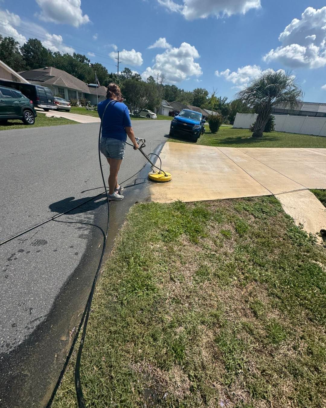Woman pressure washing a driveway on a sunny day, cleaning one section, hose in foreground.