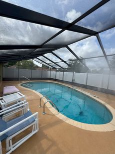 Pool under a screened enclosure with lounge chairs, surrounded by beige decking and a bright sky.