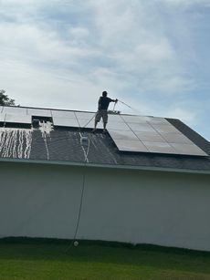 Man cleaning solar panels on a house roof with safety rope attached; sunny day.