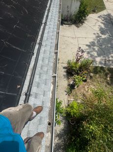 Person's feet over gutter with gravel. Building's edge, with plants and concrete on the right, black roof on the left.