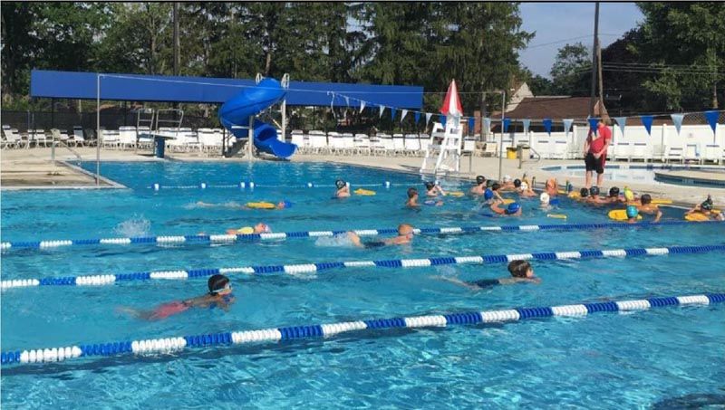 Children swimming in a blue outdoor pool with lane markers, under blue shade structure, lifeguard on duty.