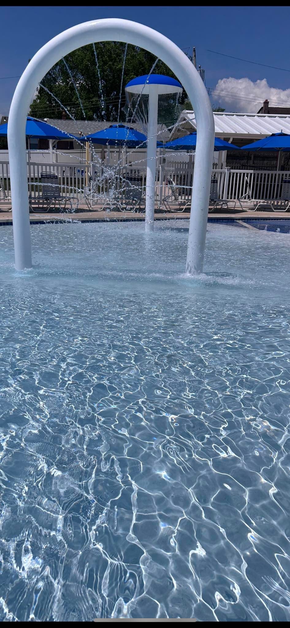A splash pad with a white arch and umbrella fountains in a pool, blue water with parasols in the background.