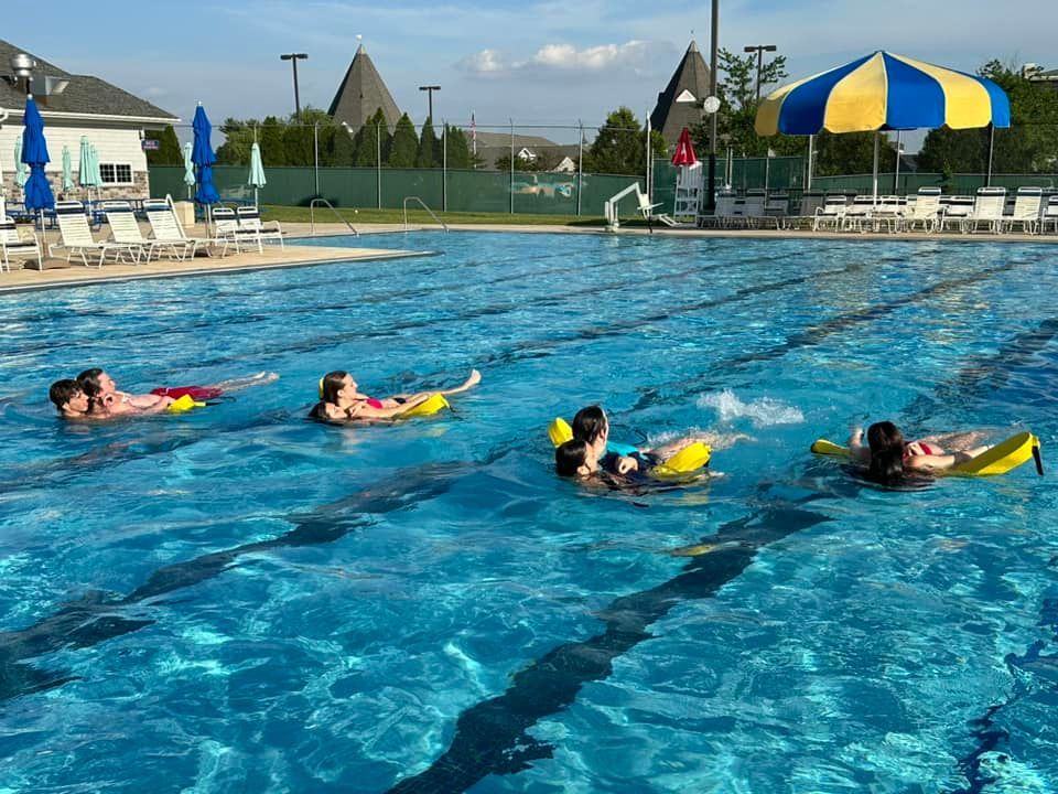 People floating on inflatable tubes in a blue pool. Yellow and red tubes are visible, with buildings in the background.