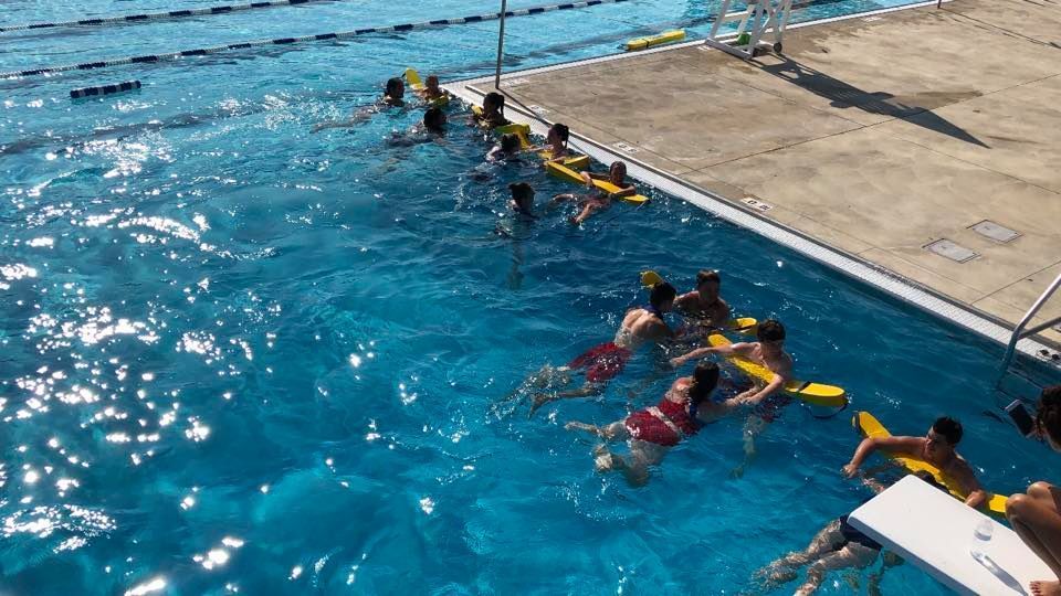 People in a pool swimming with flotation devices, under a sunny sky.
