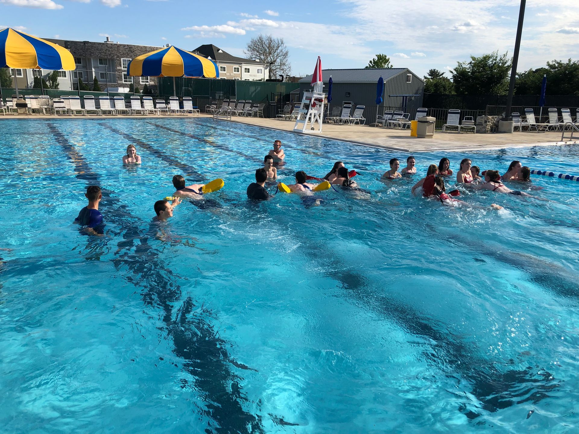 People swimming in a bright blue outdoor pool on a sunny day. Some using flotation devices.