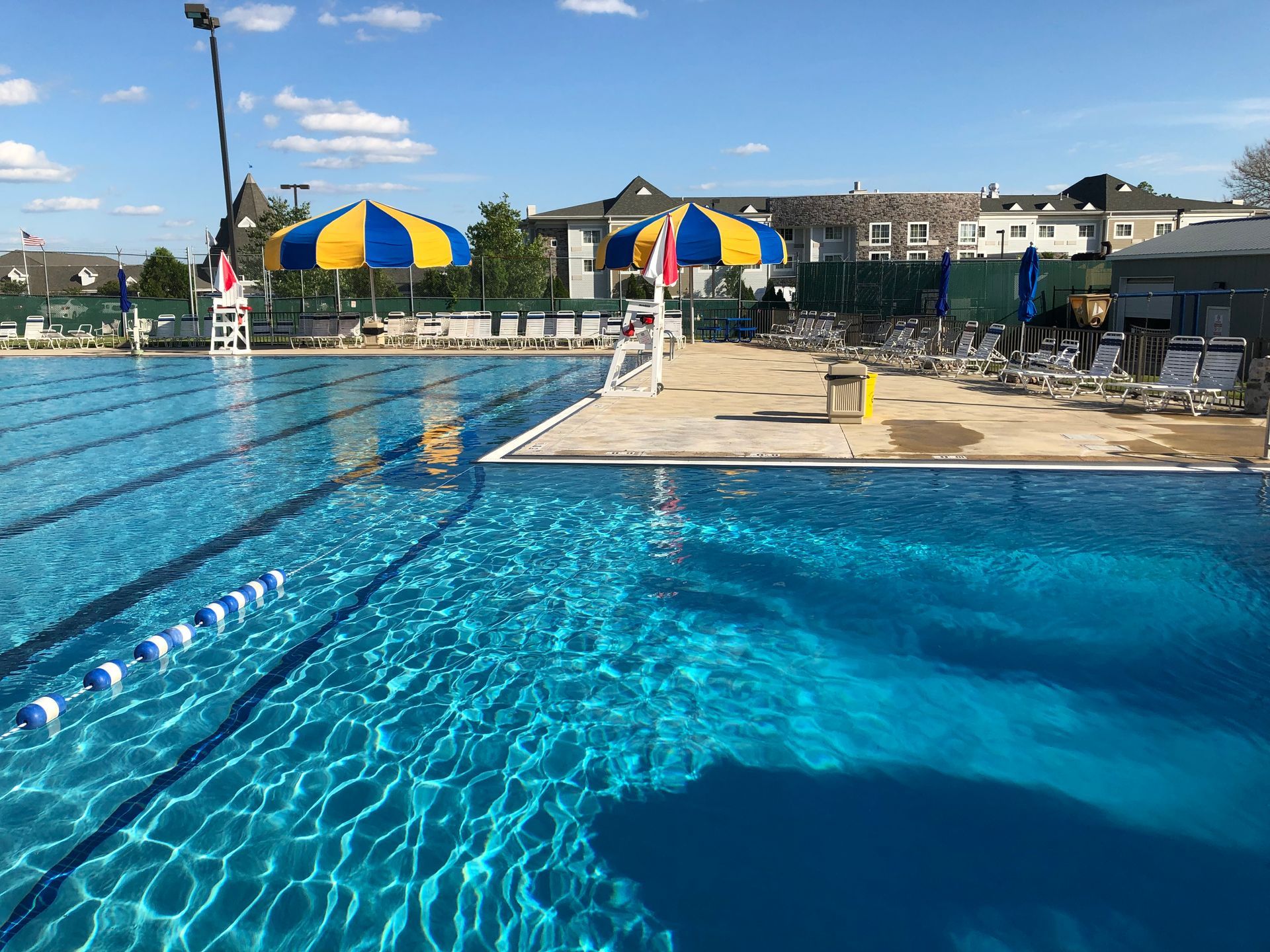 Pool with clear blue water, lane markers, sun umbrellas, and lounge chairs. Sunny outdoor setting.