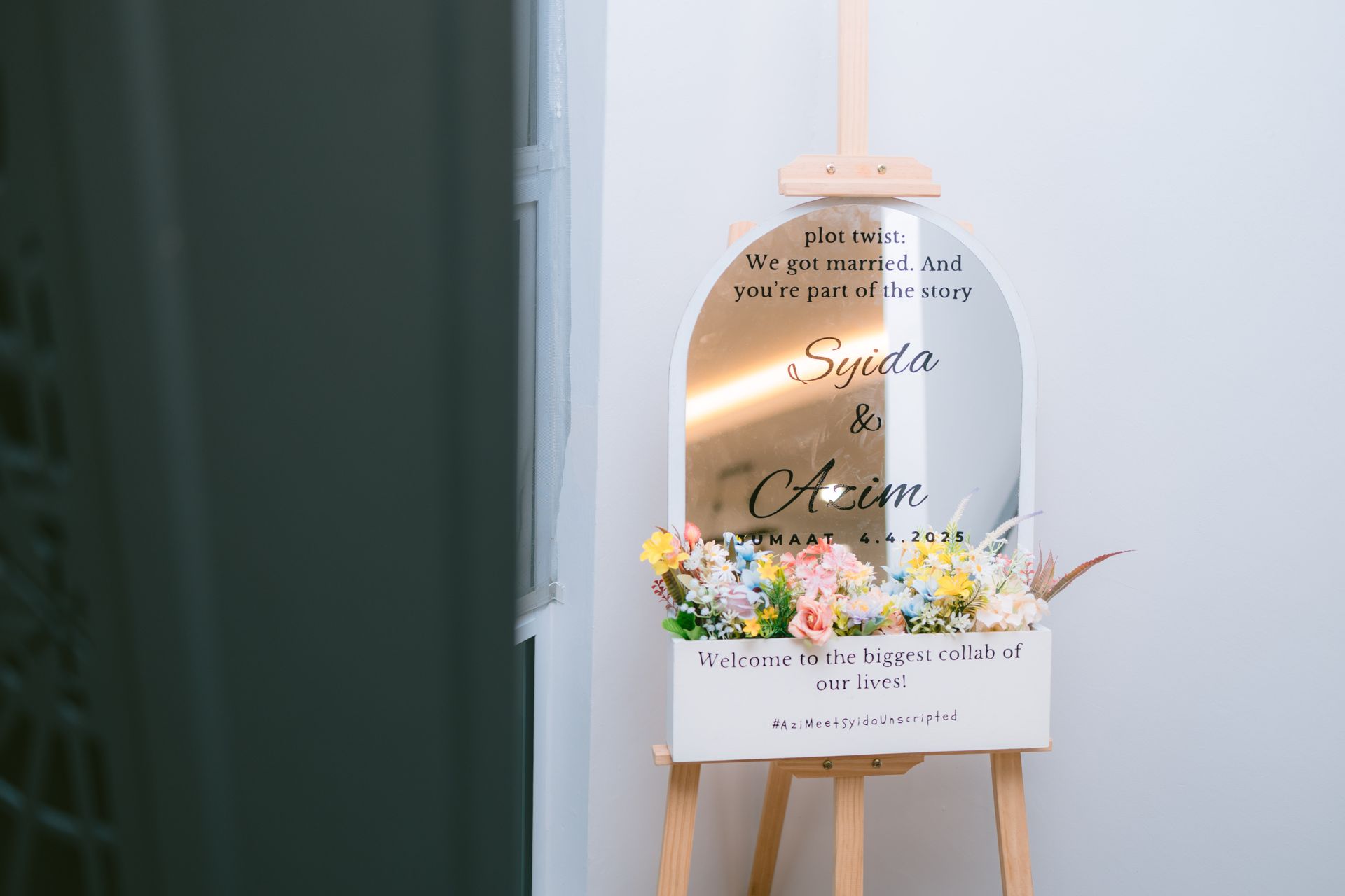 Wedding sign on an easel with flowers and a white box beneath, in soft pastel tones.