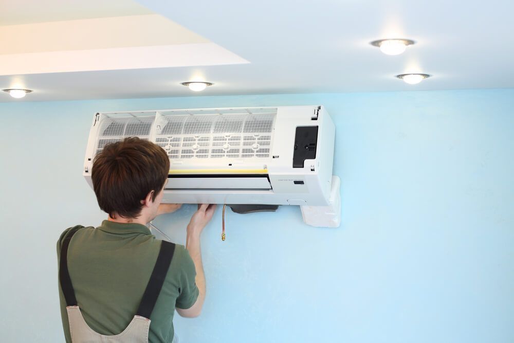 Person fixing an air conditioner mounted on a blue wall, interior, lit by ceiling lights.