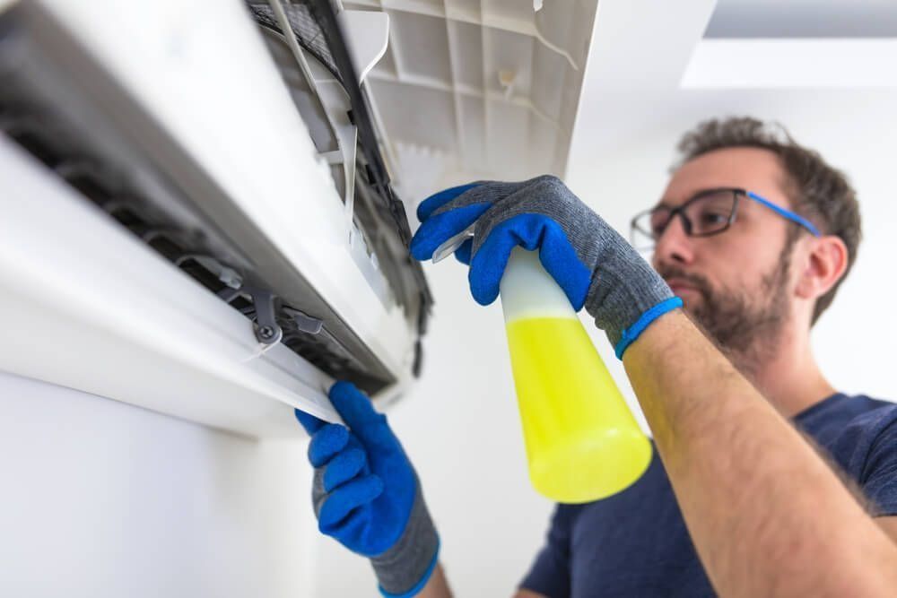 Man cleaning an air conditioning unit, spraying yellow cleaner with blue gloves.