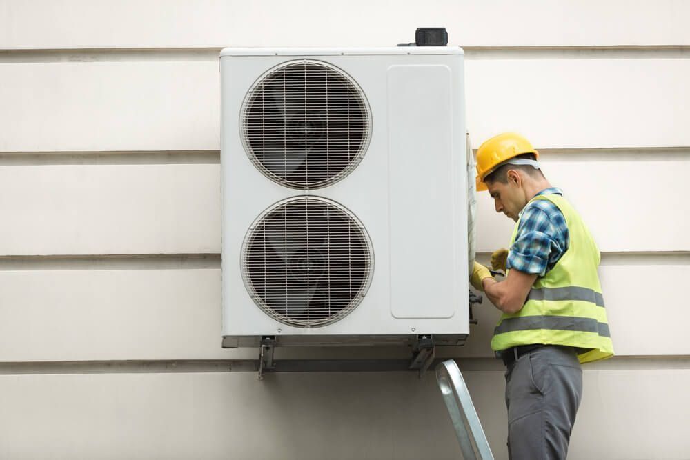 HVAC technician in yellow vest and hard hat repairs an air conditioning unit mounted on a beige wall.