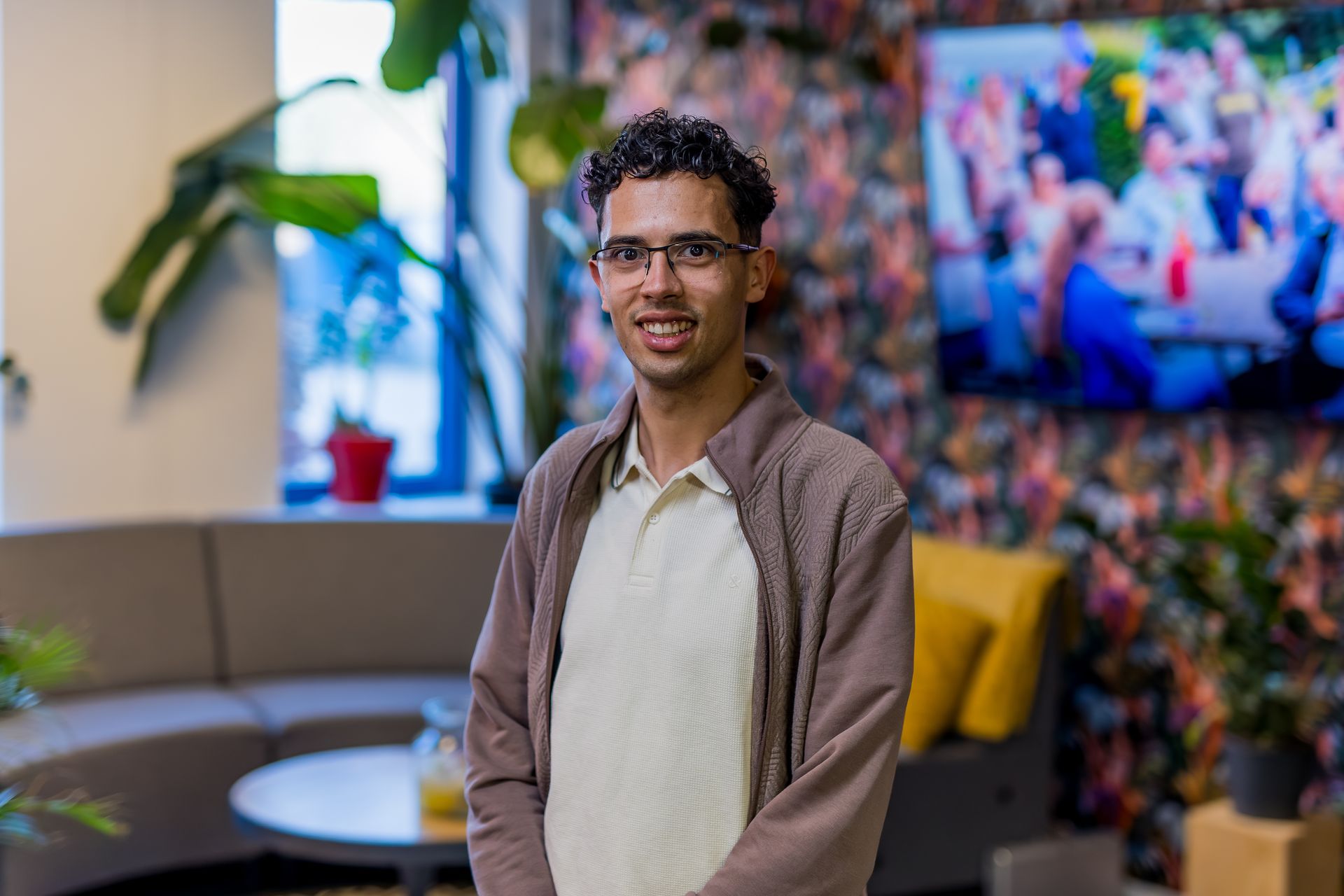 Een persoon met krullend haar en een bril, gekleed in een licht poloshirt en een bruin vest.
