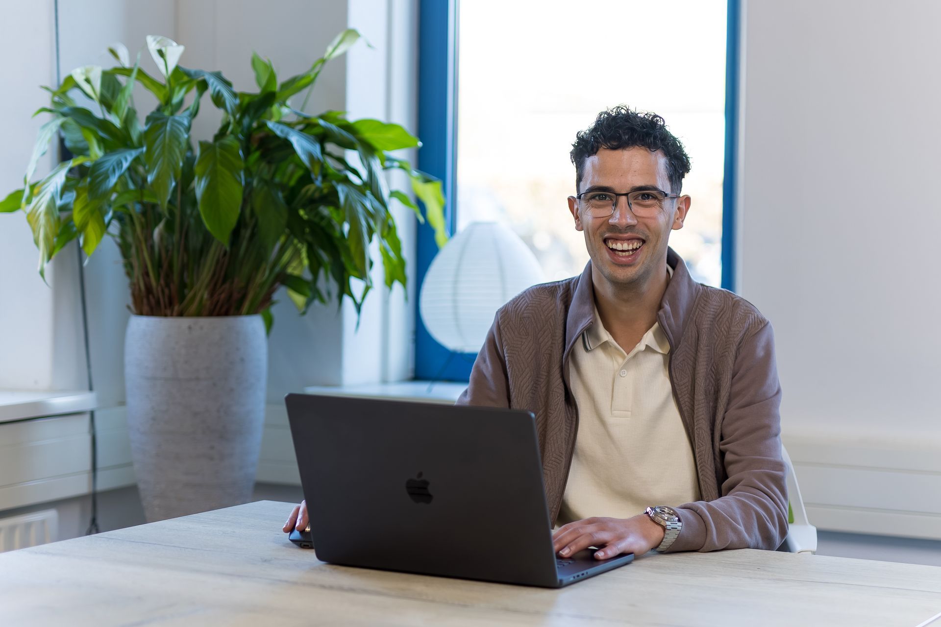 Een glimlachend persoon werkt op een laptop aan een tafel naast een grote potplant in een lichte kantoorruimte.