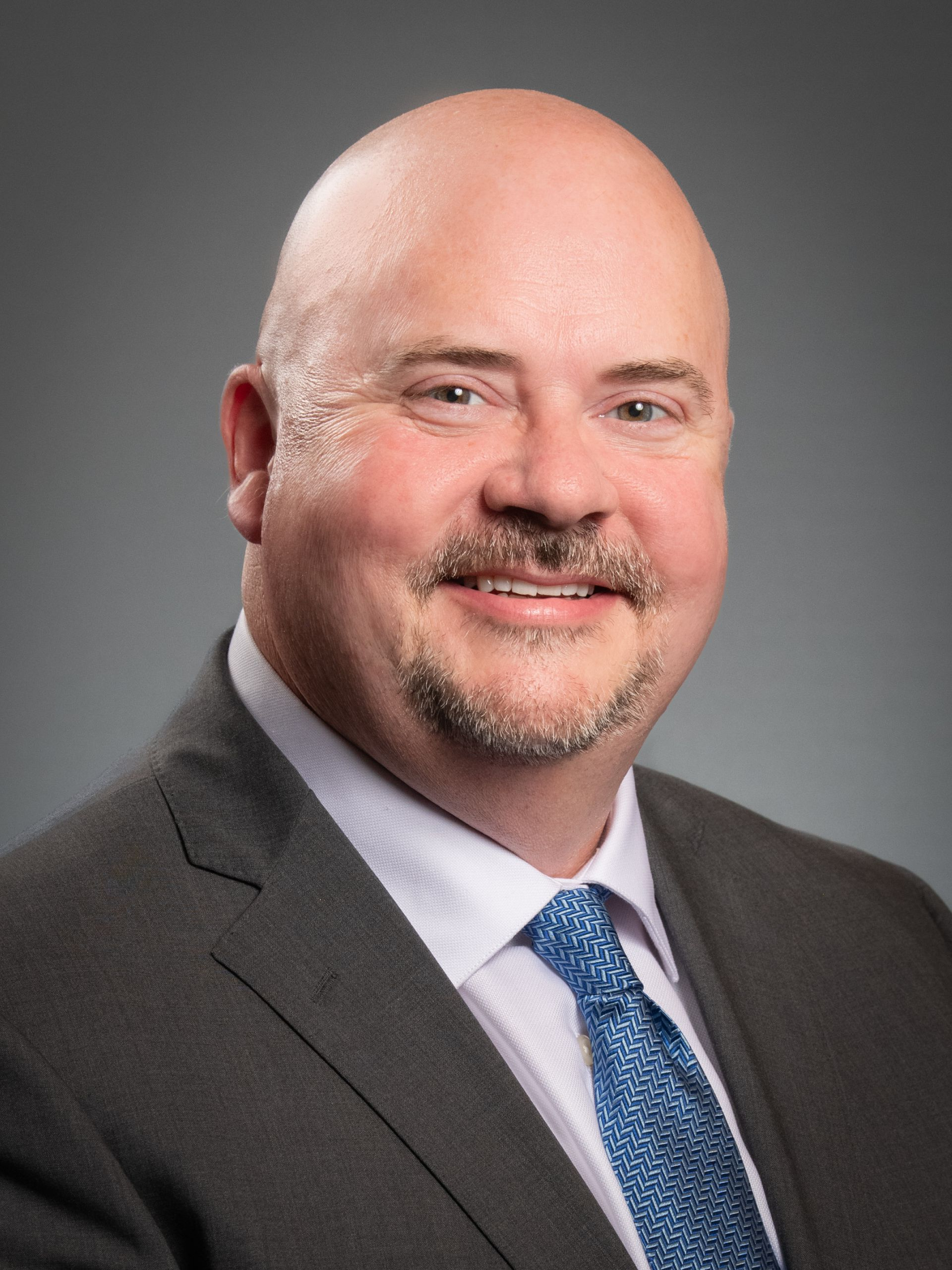 Bald man with a goatee wearing a suit and blue patterned tie, smiling in a professional headshot.