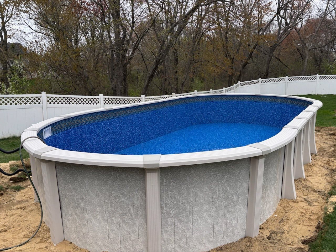 An oval swimming pool is sitting in the dirt in a backyard next to a white fence.