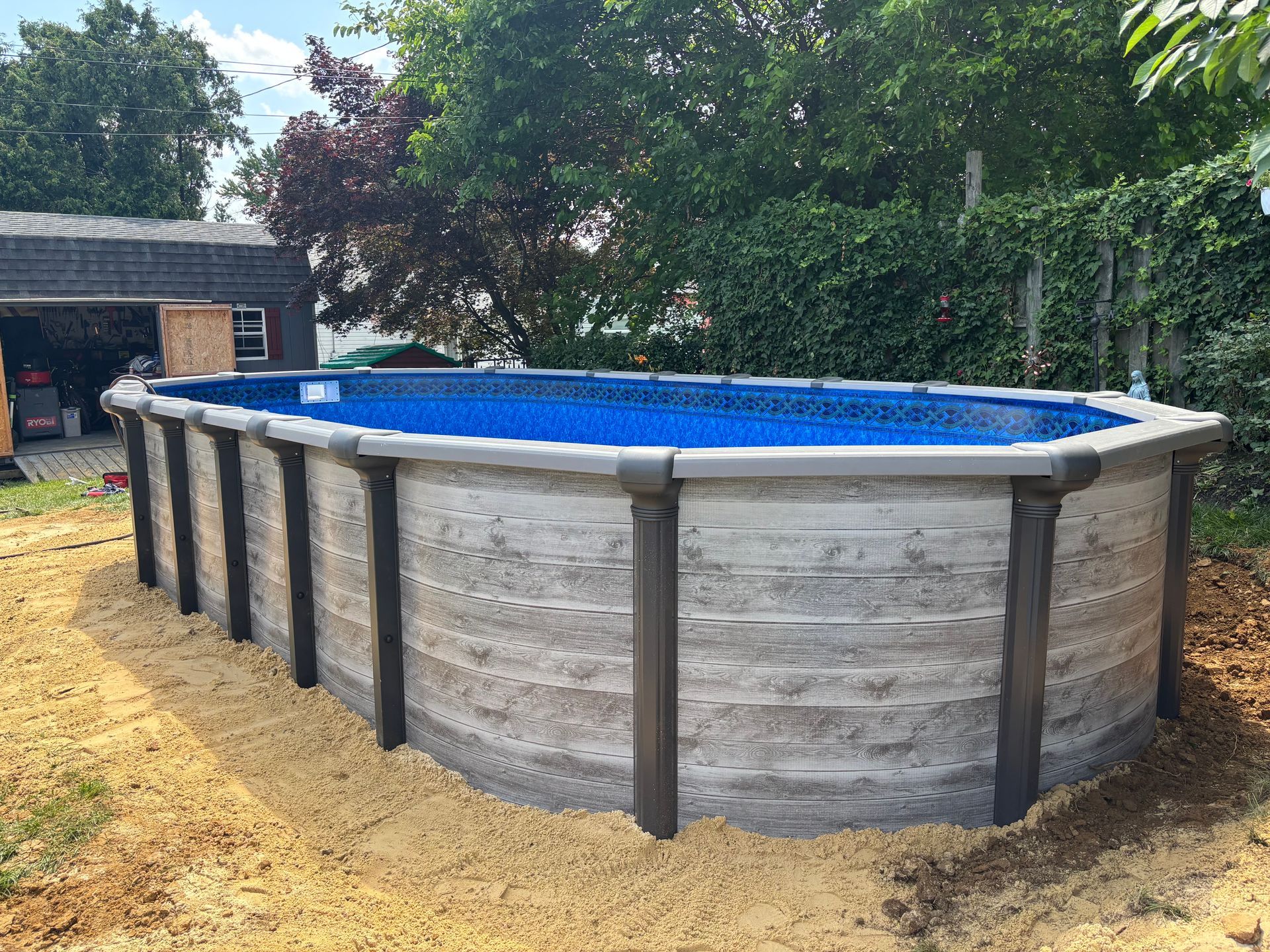 A large swimming pool is sitting in the dirt in front of a garage.
