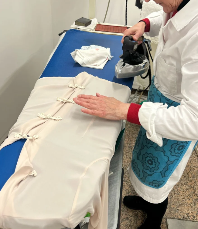 Persona che stira un top di colore chiaro su un asse da stiro rivestito di blu. Indossa un grembiule.