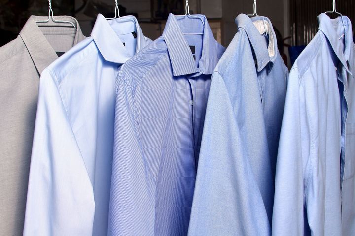 Cinque camicie abbottonate appese su un attaccapanni. Sfumature di blu e grigio.