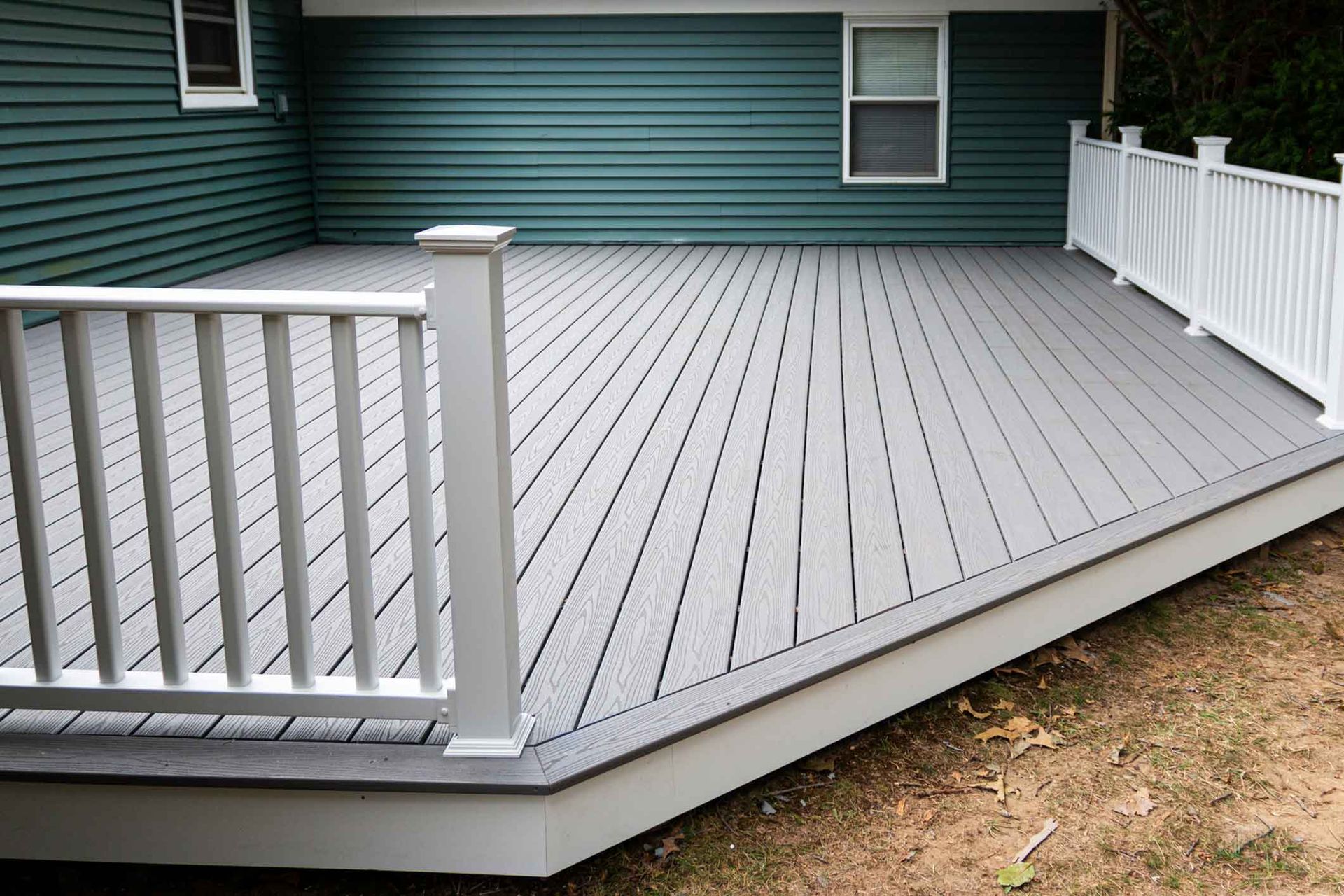 A gray deck with a white railing is in front of a green house.
